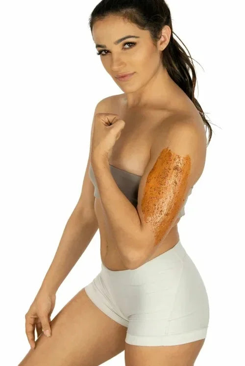 Woman applying Brightening Exfoliating Mask on her upper arm with confident expression and sporty outfit on white background