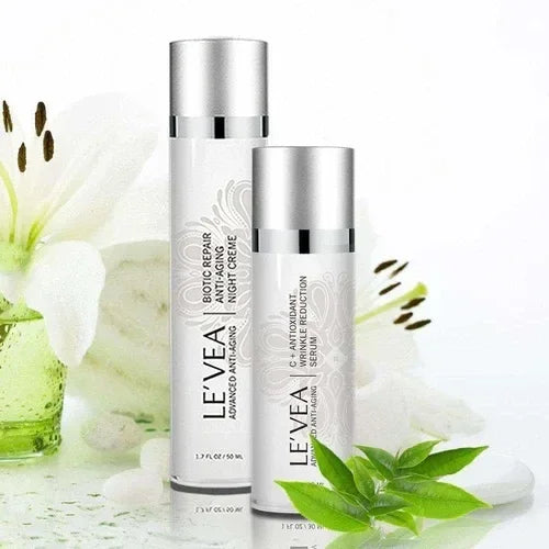 Two white skincare bottles with silver caps next to green leaves and a white flower, featuring Age Perfect: Night Repair Cream + Vitamin C+ Serum.