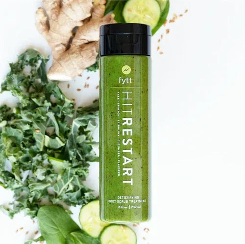 Green exfoliating scrub bottle surrounded by kale, cucumber slices, and ginger root, labeled Detoxifying Exfoliating Mask.