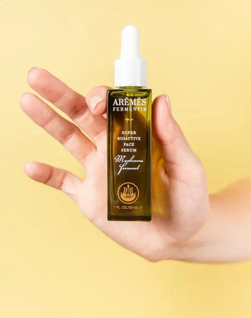 Hand holding a green glass dropper bottle of Super Bioactive Face Serum against a pale yellow background